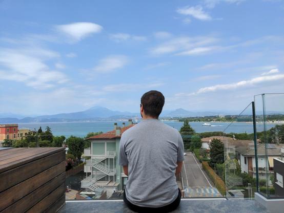 I am sitting on a rooftop terrace overlooking Lake Garda. I am seen from behind, wearing a gray t-shirt, with a scenic view of the lake, mountains, and nearby buildings under a partly cloudy sky.