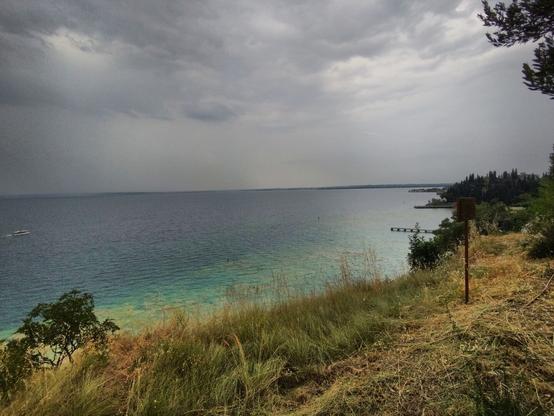 Scenic view of Lake Garda from a grassy hillside on a cloudy day. The lake displays shades of blue and turquoise, with a boat visible on the left. The hillside is covered in dry grass and scattered trees, and a metal pole stands near the edge. The sky is overcast with grey clouds.