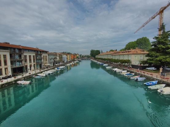 Picturesque view of a canal lined with colorful buildings and small boats on a cloudy day. The water is a clear turquoise, reflecting the surroundings. A construction crane is seen in the background, indicating ongoing development.
