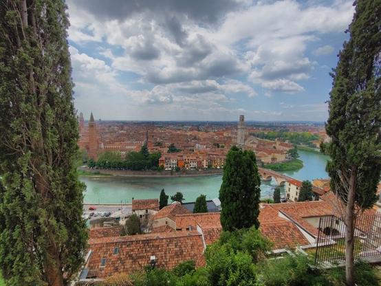 View over Verona from an elevated position. The Adige River winds through the city and a bridge is visible. The cityscape features red-tiled roofs, various towers, and spires, framed by tall trees on either side. The sky is partly cloudy, and the foreground includes rooftops and greenery.