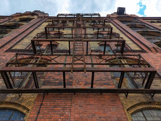 A dramatic low-angle shot looks up the imposing facade of an old, multi-storey brick warehouse building at Handelshafen in Magdeburg. The building's walls feature a pattern of dark red and lighter yellowish bricks. A prominent, multi-tiered metal fire escape, heavily rusted with a deep reddish-brown patina, extends vertically up the centre of the building. Each level of the fire escape has a narrow balcony. Numerous windows with dark frames are visible, some arched at the base and others rectangular higher up, many showing reflections of the cloudy sky. The cloudy sky is visible at the very top of the frame.