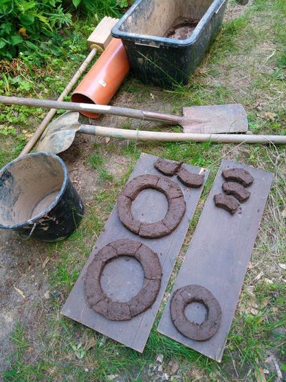 Im Vordergrund sind Kreise und Kreissegmente aus Lehmbausteinen zu sehen, in der Mitte Schaufel und Spaten, hinten eine Lehmputte.