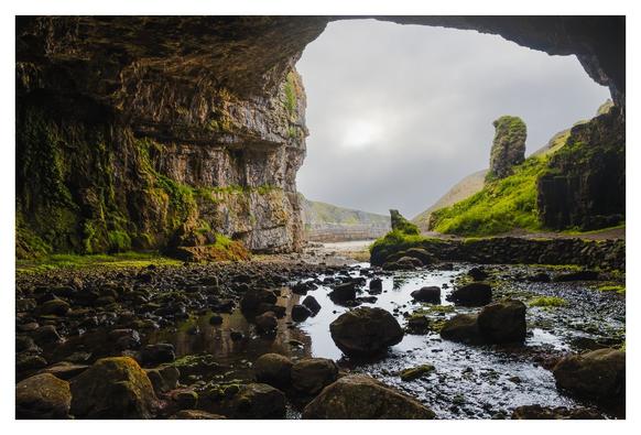 Eine beeindruckende Höhle mit üppigem Grün, die den Blick auf einen ruhigen Wasserlauf und sanfte Hügel freigibt, umhüllt von mystischem Nebel.