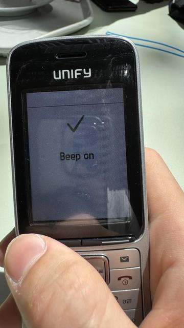 A close-up of a mobile phone displaying a screen with a green check mark and the text "Beep on." The phone is being held by a person, with a casual setting in the background.