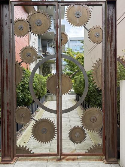 Photo of an open worked metal door composed of metal cogs of various sizes allowing the viewer to see through the door to the path beyond.  The metal frame of the door is dark brown and the cogs are tan in colour with a circular stainless steel centre.  There are green bushes on either side of the path that leads to residential buildings.
