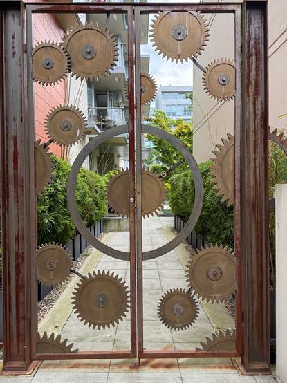 Photo of an open worked metal door composed of metal cogs of various sizes allowing the viewer to see through the door to the path beyond.  The metal frame of the door is dark brown and the cogs are tan in colour with a circular stainless steel centre.  There are green bushes on either side of the path that leads to residential buildings.