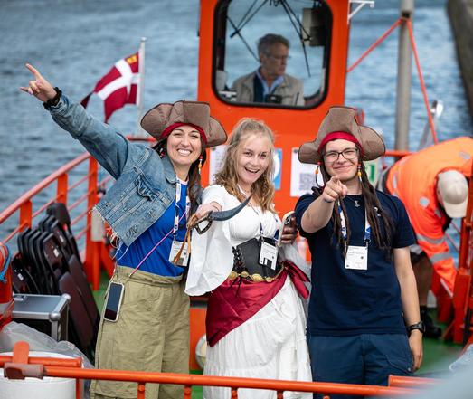 "Three people dressed as pirates smiling and pointing towards the camera aboard a boat, with a Danish flag visible in the background."