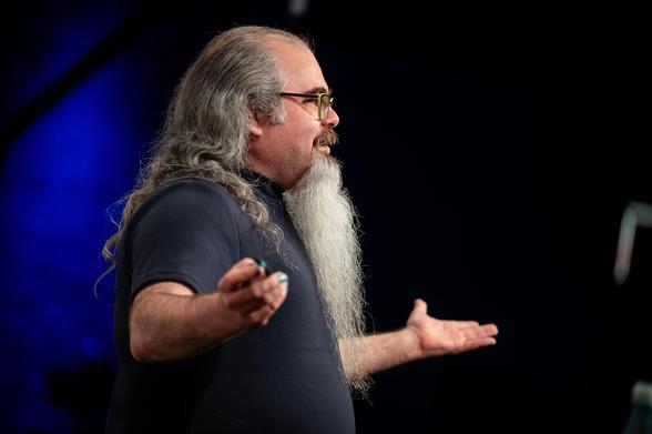"Speaker with long hair and a flowing beard presenting passionately on stage, gesturing with open hands during a talk."