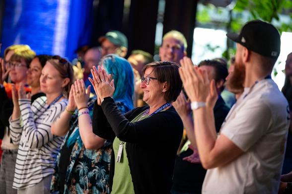 "Audience standing and clapping enthusiastically at an event, with people smiling and wearing Umbraco lanyards."