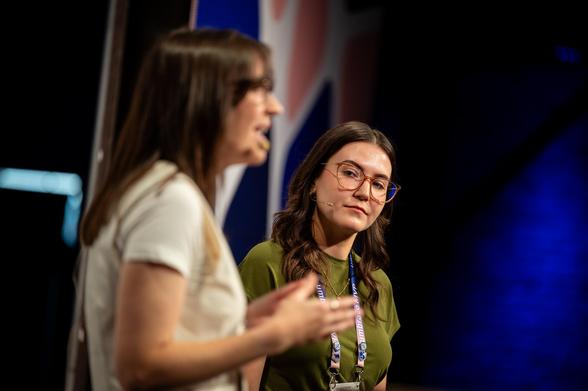"Two women on stage giving a presentation, one speaking in the foreground while the other listens attentively with a headset mic and conference lanyard."