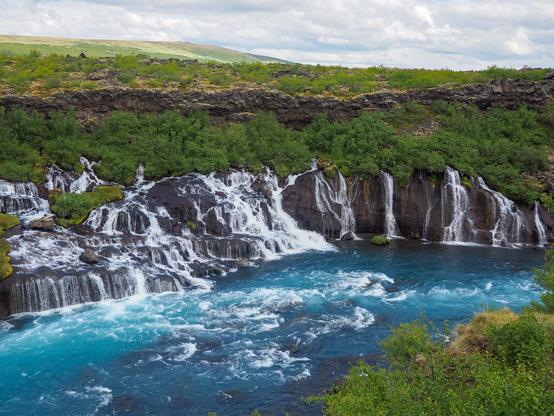 A long horizontal series of two-tiered waterfalls coming out of a lava field that's covered by moss and low bushes and falling into a blue-green river.
