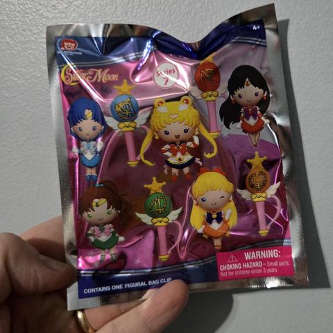 Photograph of a white person's hand holding package of a Sailor Moon dangling toy. The package has pink background with the Sailor Moon corner in the upper left-hand corner. Throughout the package it shows four different Sailor scout wands and five Sailor Scouts (Mercury, Moon, Mars, Jupiter, Venus) in a stylized toy format where they have rounded features and single solid colored dots for eyes.