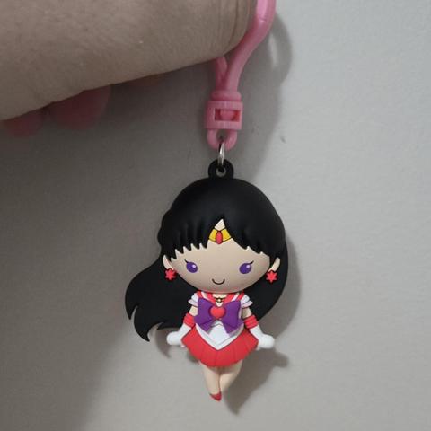 Photograph of a white person's hand holding a clip attached to a plastic Sailor Mars toy. The toy is a stylized Sailor Mars with solid purple eyes. The toy has long, black hair, and a white, purple, and red sailor scout outfit and white gloves. Her legs are crossed with red shoes visible and red earrings.