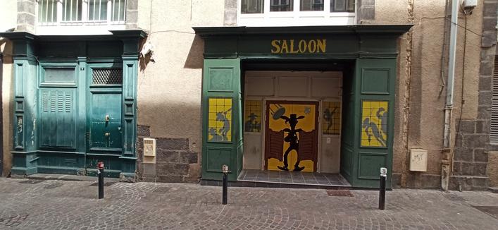 Zwei Holzverkleidungen um die Türen zweier Läden. Die linke ist hellblau/türkis gestrichen, die Rechte grün und die Schrift "Saloon" über der Tür. Auf den zwei Tür Hälften ist die Comicfigur Lucky Luke gemalt, wie er gerade die Hälften einer Saloon Tür teilt und von der Dunkelheit ins Licht tritt