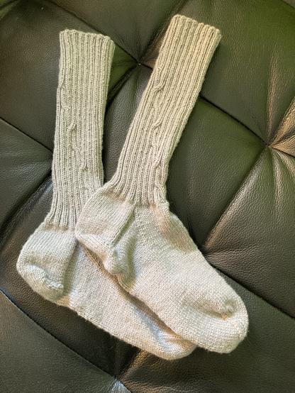 ein paar handgestrickte Wollsocken in hellgrau, mit langem Bündchen in Rippenmuster mit kleinem diagnonal verlaufendem Zopf, auf schwarzem Untergrund