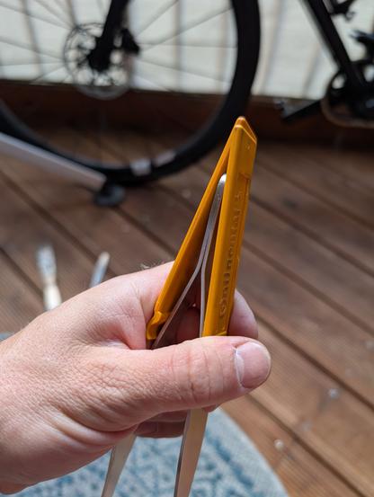 One hand holds two orange plastic tyre levers, between the two levers is a large pair of tweezers, which are conically shaped and thus spread the two tyre levers apart when pressure is applied. In the background you can see the wooden floor of a balcony and parts of a black gravel bike.