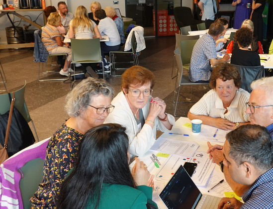 An indoor scene showing multiple small groups of people collaborating at tables. In the foreground, five older adults are actively participating in a discussion, looking at each other and shared documents.