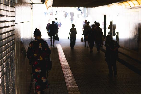 People walking through a dimly lit underground passage with light streaming in from the exit. A person in traditional Japanese attire is visible on the left, leaning against a tiled wall. Silhouettes of multiple individuals are walking toward the light in the distance. The walkway has a textured surface, and the walls have a reflective quality.