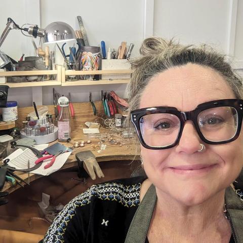 Selfie of a woman in large thick-rimmed glasses smiling at the camera. Over her shoulder can be seen a jeweller's bench that is covered in tools and partially-finished jewellery.