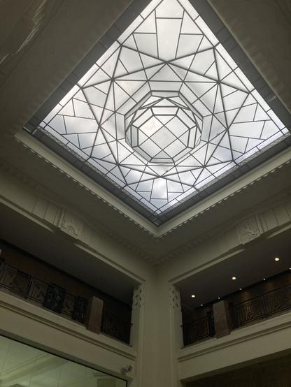 Photo d’un plafond avec une verrière art déco : un carré est empli d’autres carrés et de pièces de verre de différentes formes ; on aperçoit en bas des garde-corps