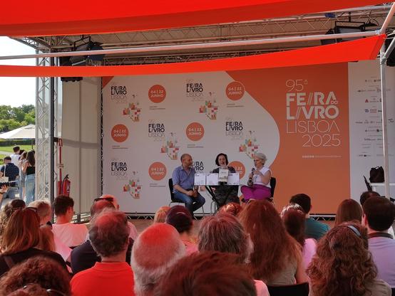 Rui Tavares, Safaa Dib e Catarina Carvalho sentados no palco da "Praça Laranja" da Feira do Livro de Lisboa. Em frente, ao palco, de costas para a câmara, o público sentado.
