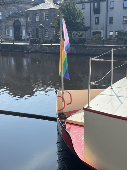 A pride flag hangs limp on a windless day, from the bow of the Ocean Mist luxury boat hotel in the Water of Leith Basin, with a moorhen on a nest on the deck just below.