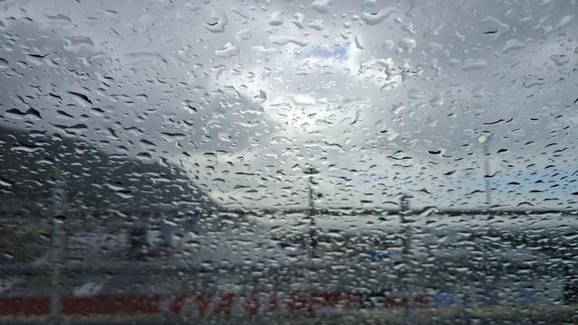 Kalk Baai Harbour seen through a rain spattered car windscreen. The view beyond the raindrops is out of focus. You can just barely see the fence in the foreground, some rooves, the grey sky, a sliver of sea and a bit of mountain.