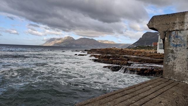 The sea outside Kalk Baai harbour over the thick concrete harbour wall. A bit of sunlight lights up the mountains in the distance, and in the foreground a grey green sea foams against rust coloured rock.
