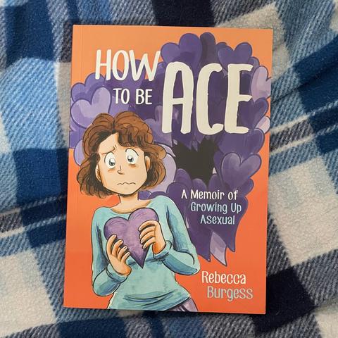 Image of a book - ‘How To Be Ace: A Memoir of Growing Up Asexual’ by Rebecca Burgess - on a blue and white blanket.