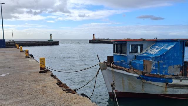 The harbour mouth at Kalk Baai from the pier. A blue and white painted fishing boat in the foreground, paintwork skuffed and faded.  The sea is grey, the sky blue between grey clouds.