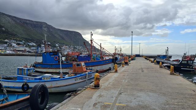 The pier at Kalk Baai harbour. Brightly painted fishing boats are moored on either side of the pier, tied to yellow bollards. The mountains in the distance are grey and green.