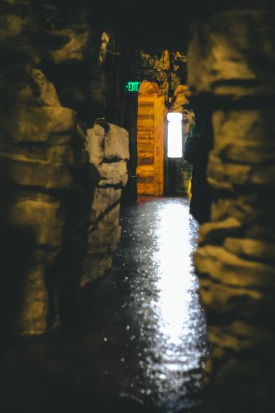 A dim, narrow stone corridor glistens with moisture under low light, leading to a warm wooden doorway glowing beneath a green exit sign, evoking a mysterious and slightly eerie atmosphere.