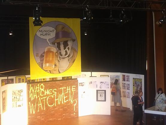 A photograph of the Dave Gibbons exhibition at this year's comic festival in Munich.
There's prints and original art by Gibbons on display, overtowered by a giant illustration of the comic character Rorschach from Watchmen, holding a German beer and mumbling "Munich? Hurm."