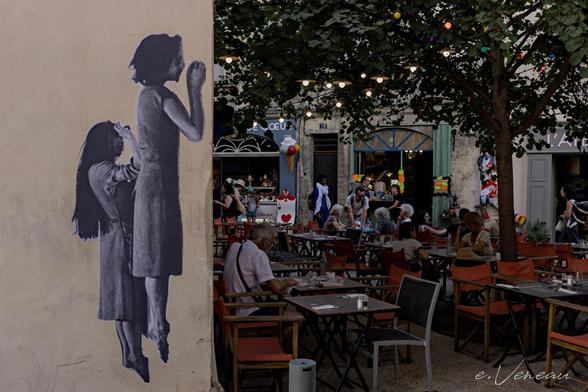 On the left is a white wall with 2 monochrome photographs of women jumping straight up in the air with their feet extended. To the right, the shaded terrace of a café, with customers and strings of lights.