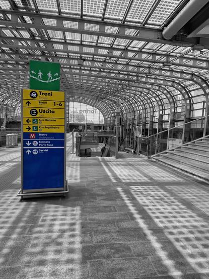 Modern train station interior with a large arched glass roof creating checkered light patterns on the floor. A colorful directional sign in the foreground provides directions for trains, exits, and metro services. Escalators and stairs are visible on the right, leading to different station levels. Sky visible through the roof, adding a sense of openness.