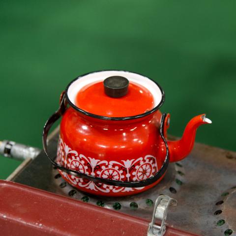 Close-up of a bright red enamel teapot with white floral patterns, white interior and black handle, sitting on top of a gas-powered camping stove.