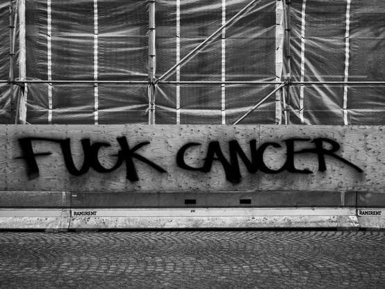 Black-and-white photograph of graffiti on a plywood construction barrier, reading "Fuck Cancer" in very large, bold black letters. The background features scaffolding and covered frames.