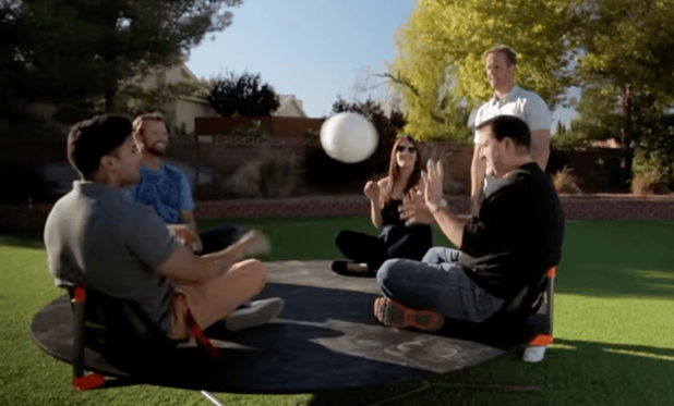 Four people sit on a circular platform in a park setting as a white ball is captured mid-flight between them during a Coriolis Effect physics demonstration.