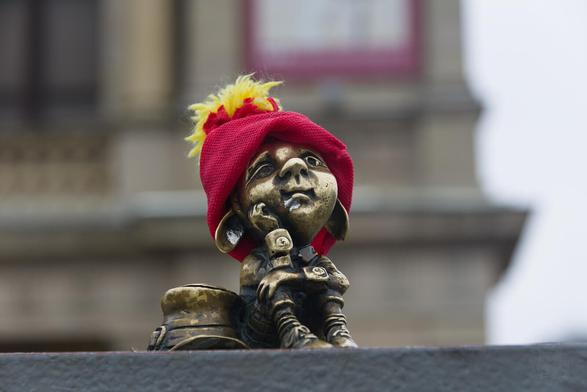 The image captures a palm-size statue of a man sitting on the ground. The man, crafted from metal with hues reminiscent of bronze or brass, appears to be thinking or daydreaming with his arms resting on his raised knees, and his left palm supporting his head. He has a big head and big slightly pointy ears drooping down. He is dressed in a traditional outfit complete with long-sleeved shirt, checked  trousers and shoes with large buckles. His large hat, resembling to Leprechaun's one, is placed on the ground to his right side. The figure is adorned with a hand-made red hat that sports yellow feathers at the top, adding a touch of whimsy to its otherwise serious demeanor.

The statue stands out against a blurred backdrop featuring architectural elements such as windows and balconies, suggesting an urban setting for this tableau. The focus on the figure suggests it might be a point of interest or landmark within its location.