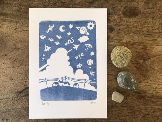 Linocut print of a tony village under a big blue sky filles with flying objects. The picture is printed in deep blue ink.