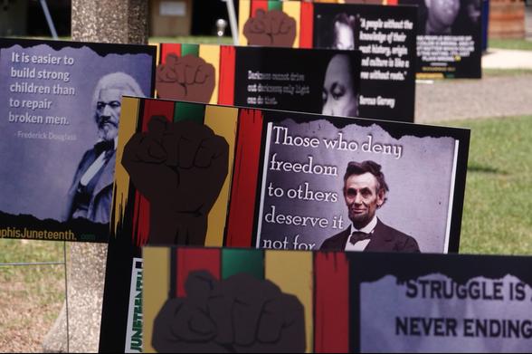 A collection of Juneteenth signs displayed on a grassy area. Prominent among them are historical quotes with portraits, including Frederick Douglass (“It is easier to build strong children than to repair broken men”) and Abraham Lincoln (“Those who deny freedom to others deserve it not for themselves”). Several signs feature the raised fist symbol and Pan-African colors (red, black, green, and yellow), symbolizing Black liberation and resistance.
