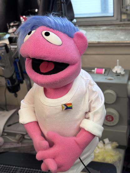 Human hand puppet with pink fleece, blue hair and white shirt wearing a Pride flag pin, left view