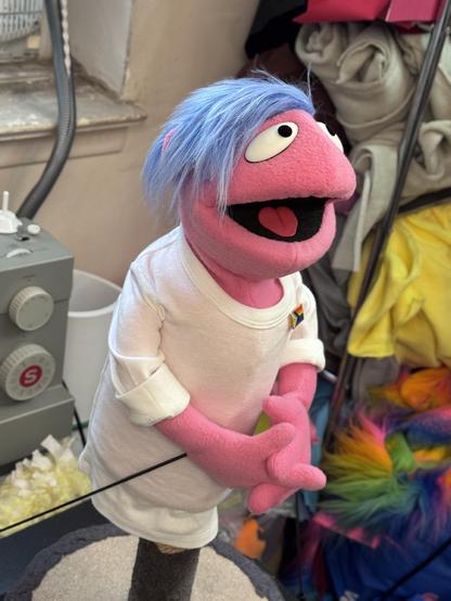 Human hand puppet with pink fleece, blue hair and white shirt wearing a Pride flag pin, right view