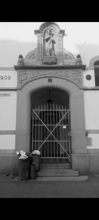 A large doorway with a gate and inside that is a outdoor hallway with a hanging lamppost and a smaller door that leads inside the actual building. Outside the first door is a small staircase and some flowerpots on the left side. The doorway is looking like its framed because of how the building was built and above it it is decorated with a figurine stanind on a shelve. The text between the door and the figurine reads bathhouse.