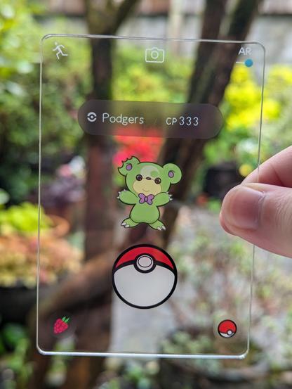 photo of a transparent acrylic card made to look like a Pokemon Go catching screen, featuring a green Teddiursa with a bow-tie and a damaged ear as the Pokemon being caught