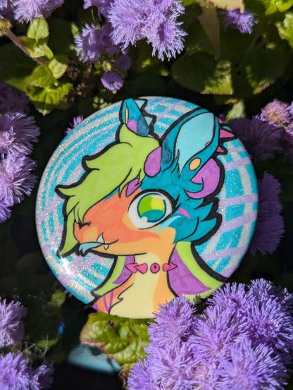 photo of a pinback button badge featuring a drawing of a very colourful anthro animal on a background of abstract colours and glitter