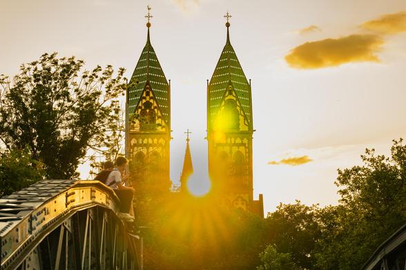 Three people sit on the edge of a metal bridge, silhouetted against a vibrant sunset. They look towards twin church towers with green-tiled roofs and intricate Gothic architecture. The sun appears between the towers, creating a stunning halo effect and casting a warm glow. Trees frame the scene, and the sky is lit with soft, golden hues.