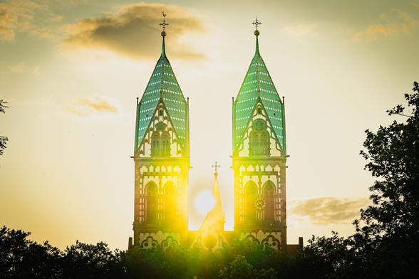 A bright sunset illuminates twin church towers with green-tiled roofs and intricate architectural details. The sun sits directly between the towers, creating a glowing halo effect. Silhouetted trees frame the scene on both sides, and a partly cloudy sky is tinged with warm golden hues.