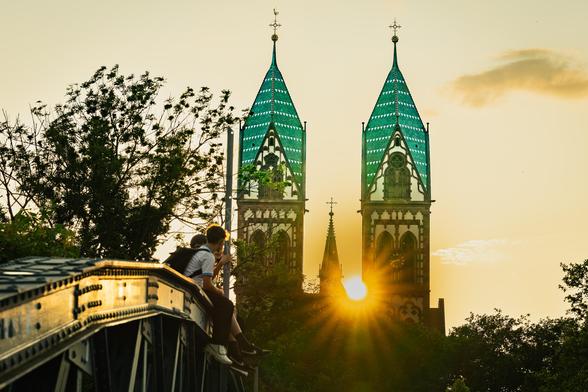 A bright sunset casts a warm glow behind twin church towers with green-tiled roofs and ornate details. The sun is positioned between the towers, creating a halo effect. In the foreground, two people sit on the edge of a metal bridge, looking toward the horizon. Silhouetted trees frame the scene, with the sky displaying soft clouds and golden hues.