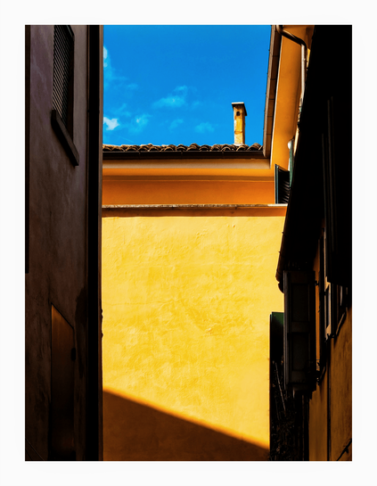 This photo is of the upper portion of a narrow alleyway between two buildings with a building at the end. The buildings on the left and right are in the shade and are a dark brown colour. The other at the end is a bright, sunny yellow. The yellow building has a tiled roof and a chimney visible against a blue sky. There are a few whispy clouds on the left. On the shaded buildings there are windows and shutters visible.